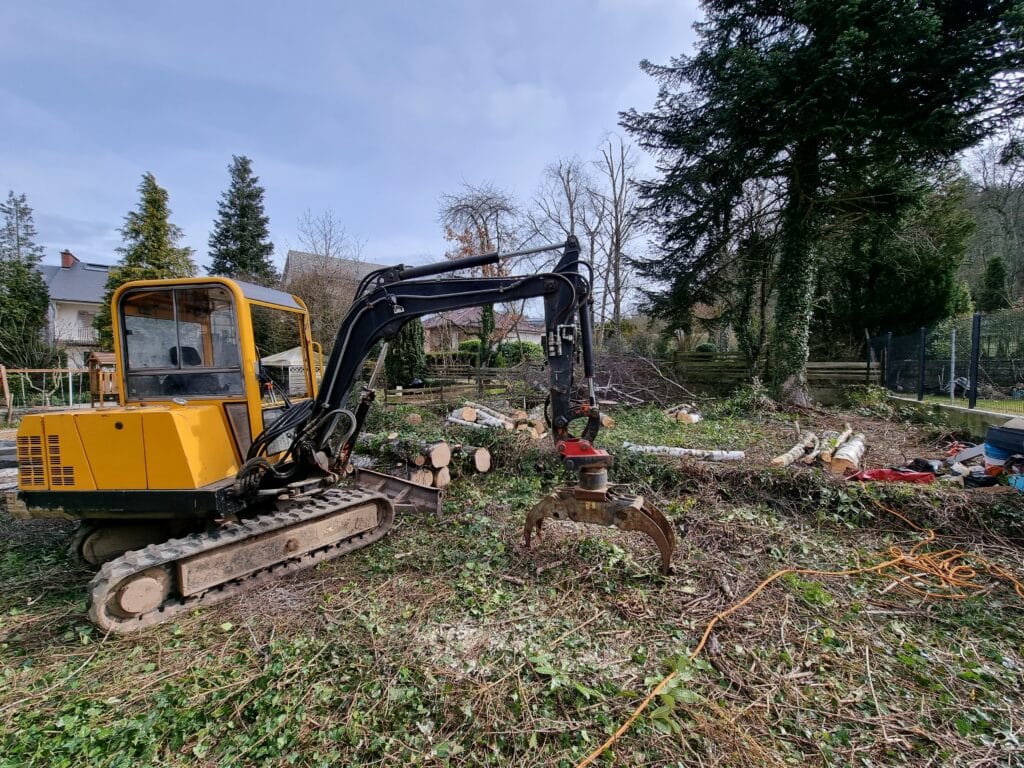 Une mini-pelle avec un grappin à bois utilisée chez un particulier pour débarrasser des troncs d'arbre.