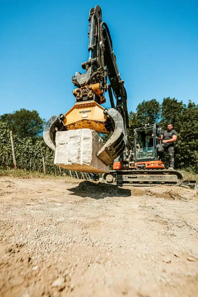 Bagger beim Transport eines Natursteinblocks für den Bau einer Mauer.